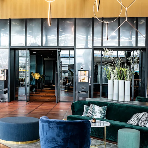 Luksuriøs hotellobby med bløde siddepladser og elegant indretning. Store glasdøre åbnes til et stilfuldt interiør med planter og stemningsbelysning, hvilket skaber en sofistikeret og indbydende atmosfære.