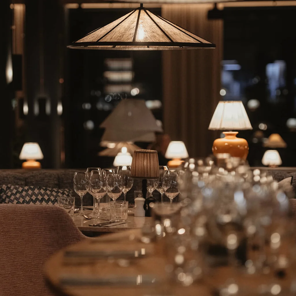 Bord dekket med vinglass i en svakt opplyst restaurant. Myk belysning fra lamper skaper en koselig atmosfære, med polstrede seter og dekorative detaljer som forsterker settingen.