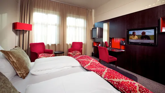 Et moderne hotellrom med en seng, røde stoler, et skrivebord, og en TV som viser to menn.