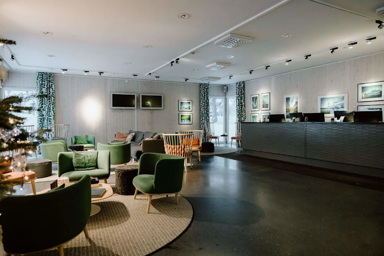 Reception område med grønne stole omkring borde, ved siden af en moderne trædisk med computerskærme. Væggene har indrammet kunst i et lyst, rumligt miljø.