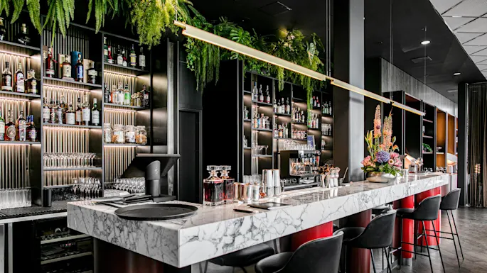 A marble bar counter with black chairs displays cocktail-making tools and a flower arrangement. Shelves behind hold various liquor bottles amid ambient lighting and lush hanging plants.