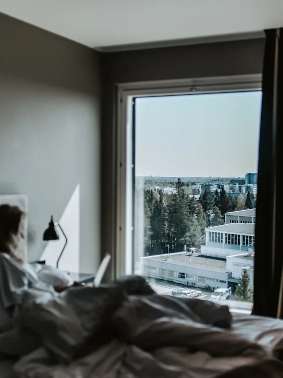 Någon i sängen med en laptop, tittar ut genom ett fönster mot en avlägsen stad och träd en solig dag.