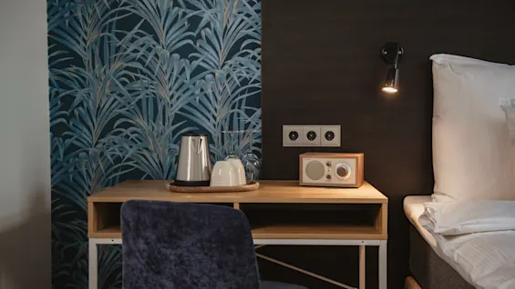 Desk holds kettle, glass, and radio; positioned beside a bed with a reading light. Blue patterned wallpaper contrasts with dark accent wall in a cozy, modern bedroom.