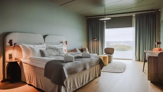 En sirligt redd seng med puder og håndklæder står i et moderne hotelværelse, med et skrivebord og stol, blød belysning og et stort vindue med flydende gardiner med udsigt til en naturskøn udsigt.