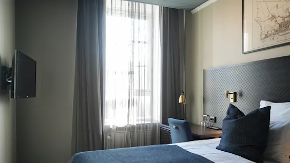 En seng med blått sengetøy og puter er ved et vindu med tynne gardiner. En veggmontert TV er motsatt, og et skrivebord med lampe er i nærheten i rommet.