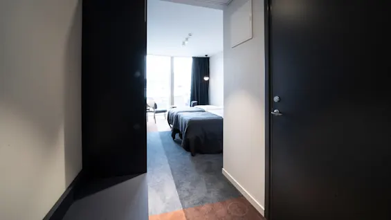 A minimalistic hotel room features a neatly made bed with dark covers, close to large windows. The room contains simple furniture and is illuminated by ceiling lights.