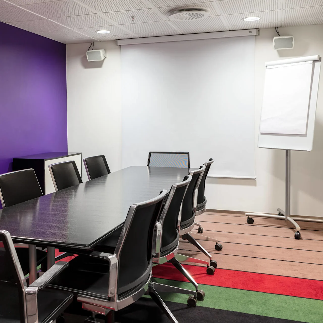 A long conference table with black chairs is surrounded by a purple and white room. A flip chart and a blank projector screen are set up at the front.