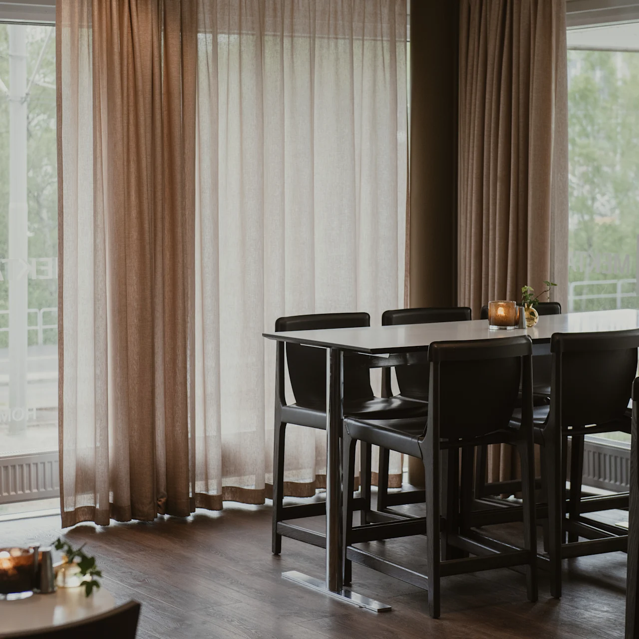 High wooden tables and chairs stand in a softly lit cafe beside sheer curtains, with trees visible outside large windows. A small candle sits on the table. Text on the window reads: "INTEGRACEN".