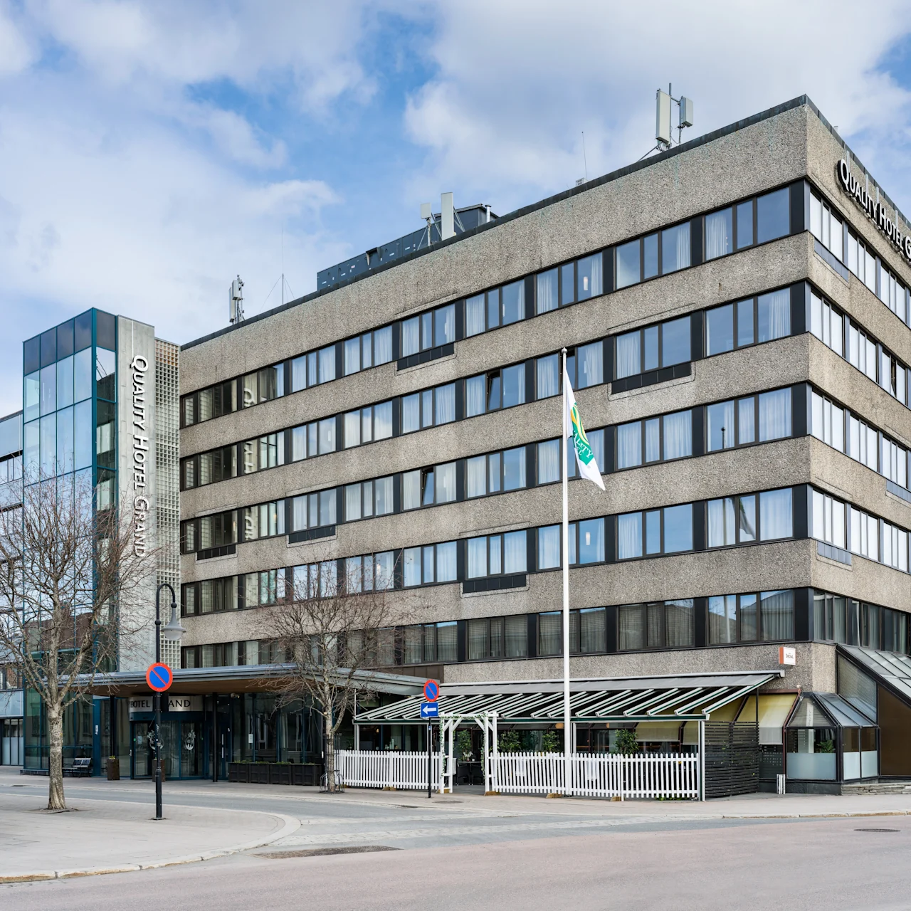 A multi-story hotel building stands quietly on a city street; its sign reads "Quality Hotel Maritim," with flags fluttering nearby under a blue sky.