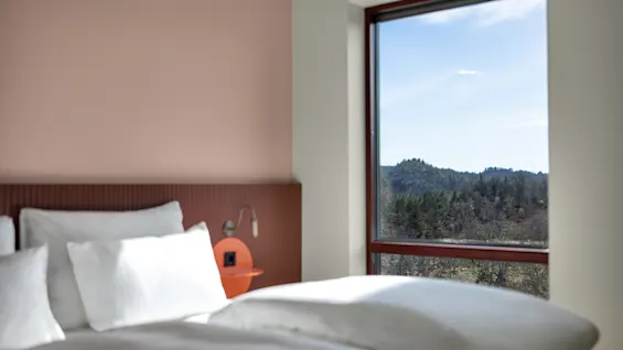 Et komfortabelt hotellrom med en seng og et stort vindu som tilbyr en rolig utsikt over en frodig grønn skog og åser.