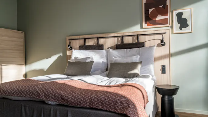 En pent oppredd seng med hvite puter og et mønstret brunt teppe. Den står mot en lysegrønn vegg med abstrakt kunst. Sollys strømmer inn gjennom et vindu.