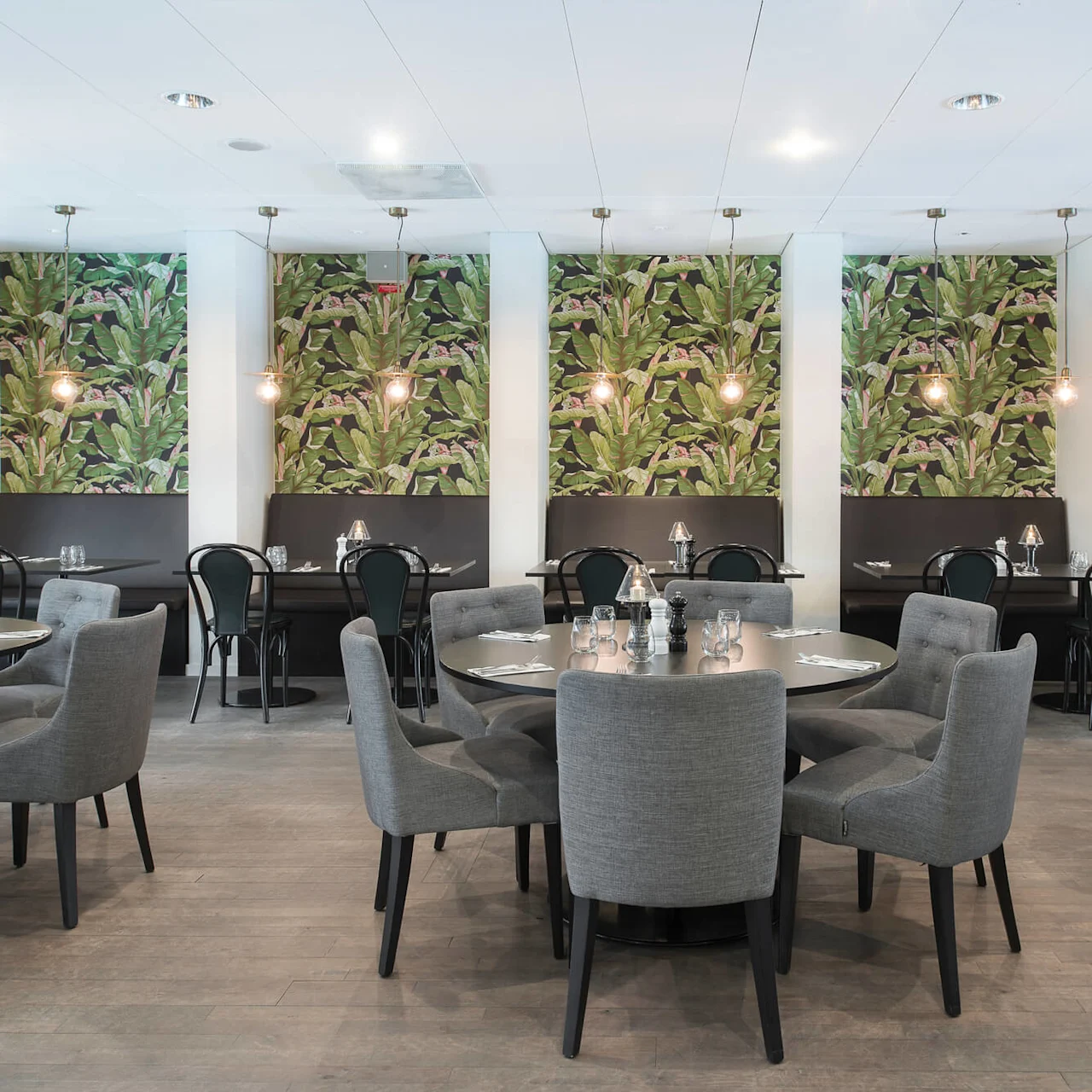 Runda bord omgivna av grå stolar är placerade i en restaurang med tropiskt bladmönstrad tapet, hänglampor och svarta bås, vilket skapar en modern matmiljö.
