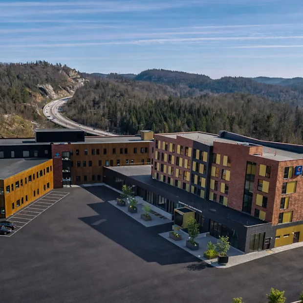 Ett modernt hotellkomplex med parkering, omgivet av skogsklädda kullar och en motorväg i bakgrunden. Synlig text: Hotell.