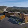 Ett modernt hotellkomplex med parkering, omgivet av skogsklädda kullar och en motorväg i bakgrunden. Synlig text: Hotell.