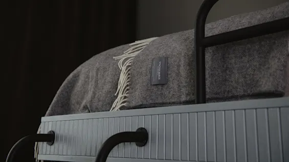 A gray woolen blanket rests folded on a blue upper bunk bed with black metal bars. Visible label reads "STACKELBERGS SWEDEN."