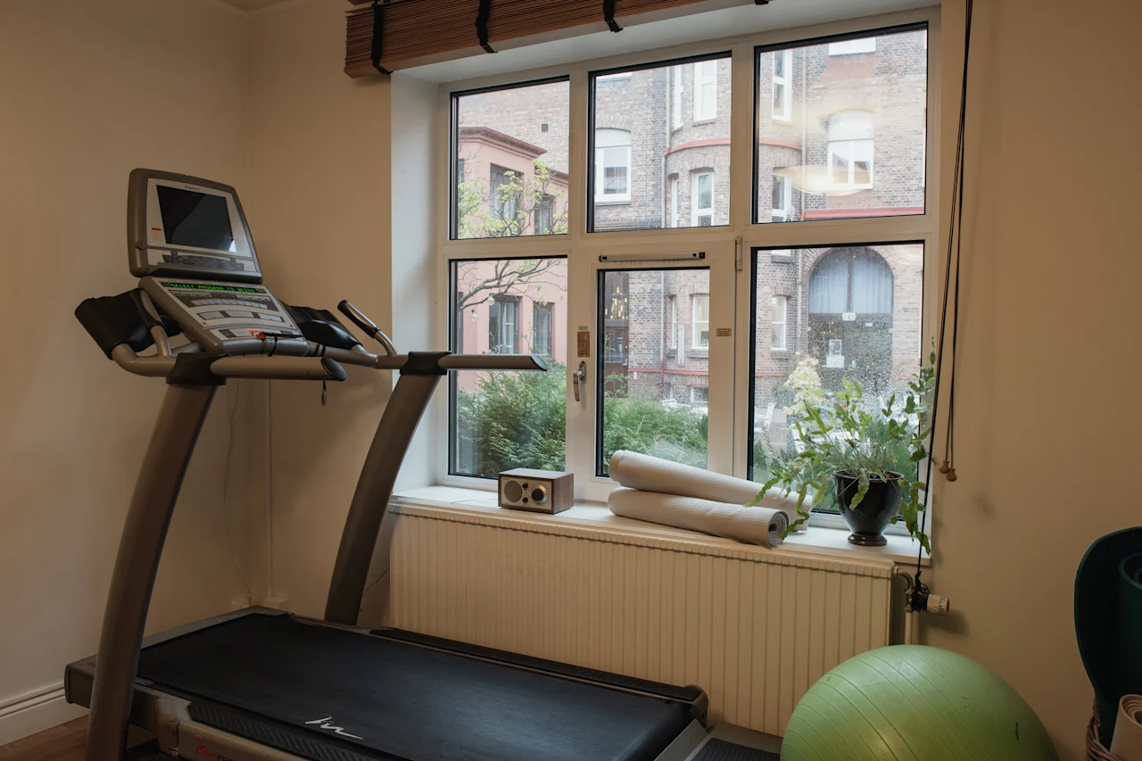 An indoor fitness area with a treadmill by a window, offering a view of urban buildings and greenery.