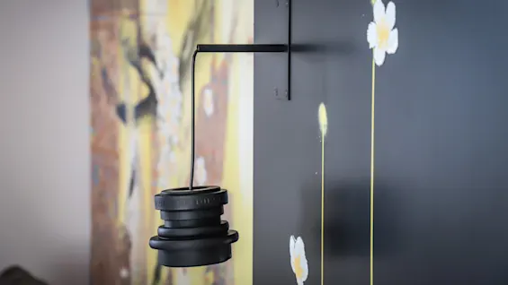 A black, industrial-style pendant light fixture hangs from a bracket on a wall, which is decorated with white flowers. A blurred, abstract painting is visible in the background.