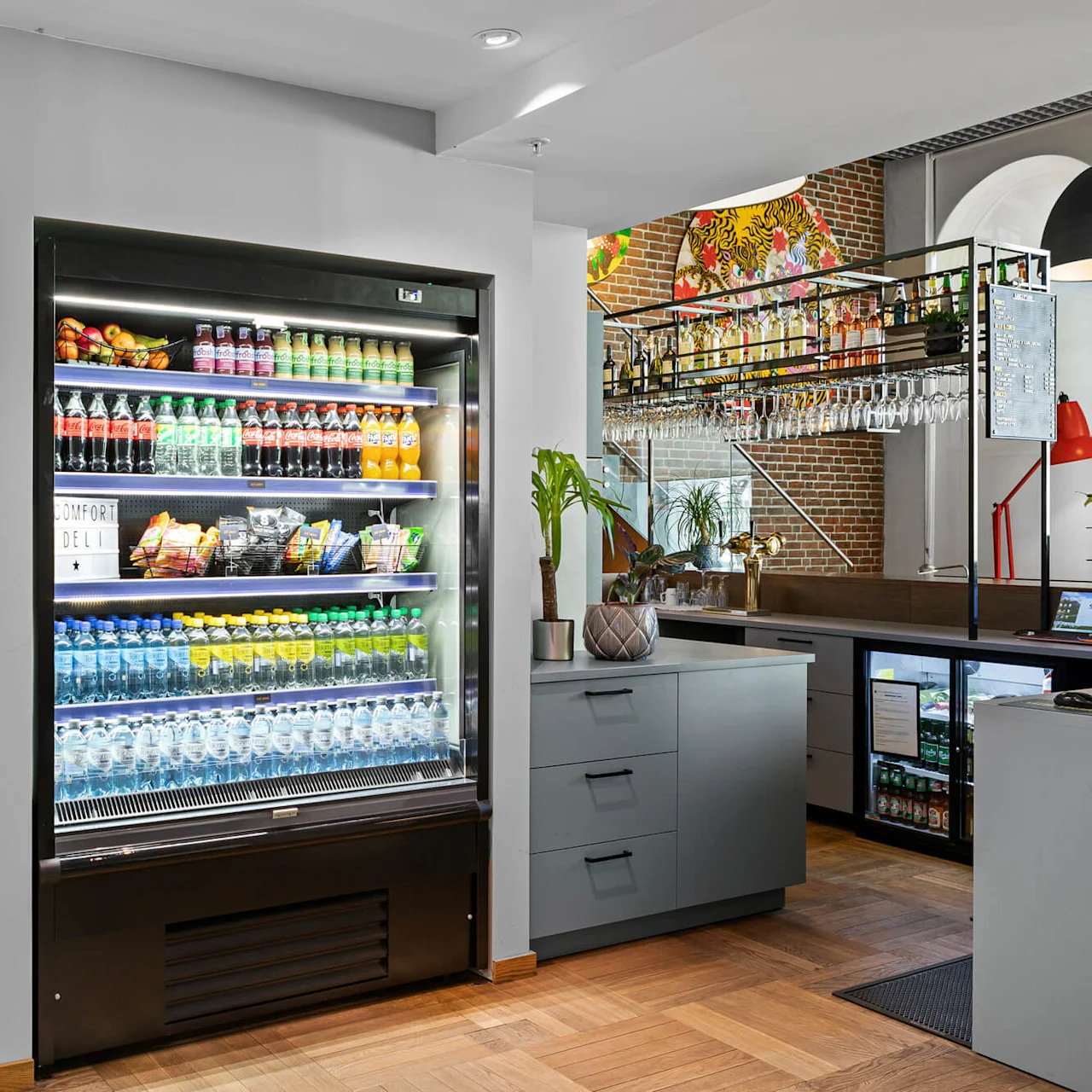 Refrigerator displays drinks in a modern cafe. Shelves hold a variety of beverages like water and soda. Nearby, snack racks and umbrellas are organized against a wall. "Comfort Deli" sign visible. 