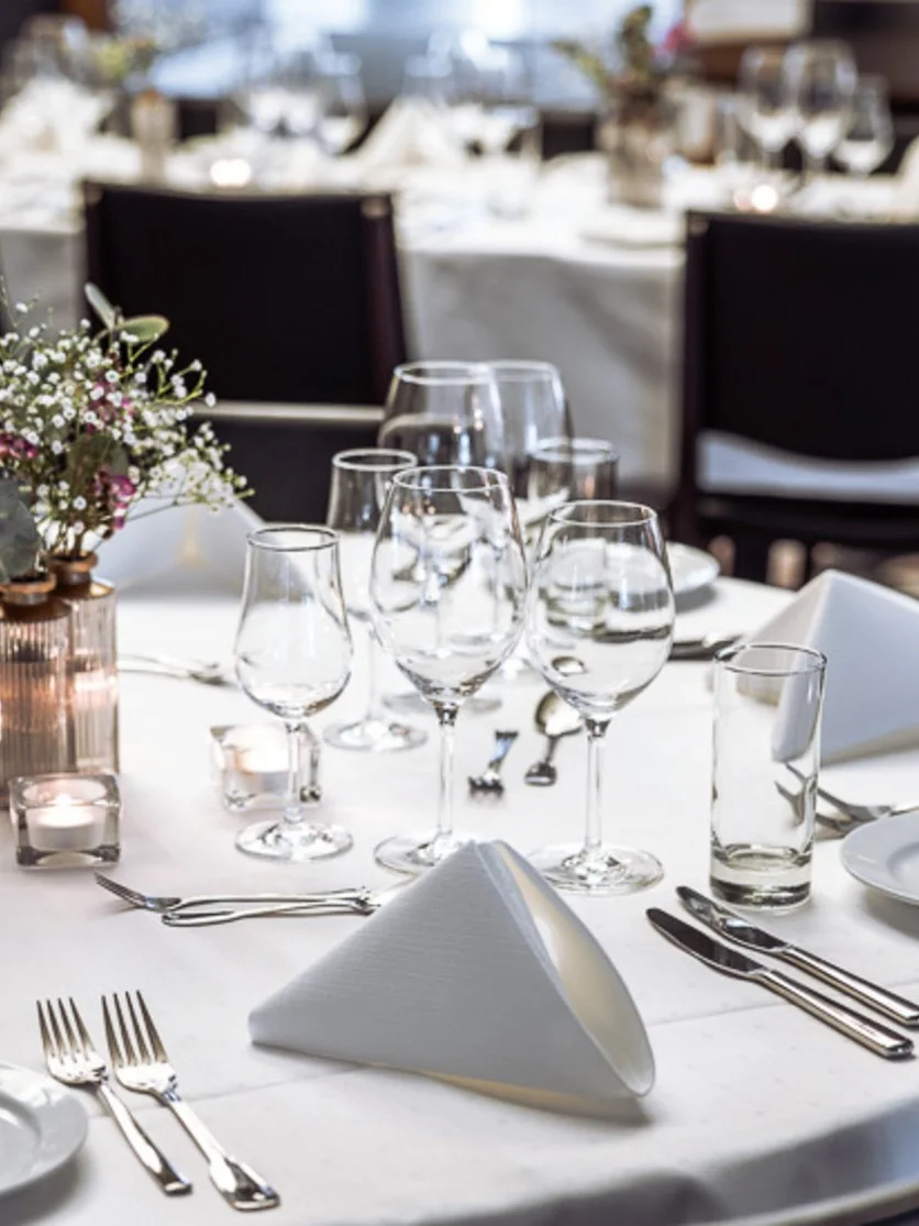 A round dining table is elegantly set with white linens, folded napkins, cutlery, glassware, and a small floral centerpiece; surrounded by neatly arranged chairs in a formal dining room.