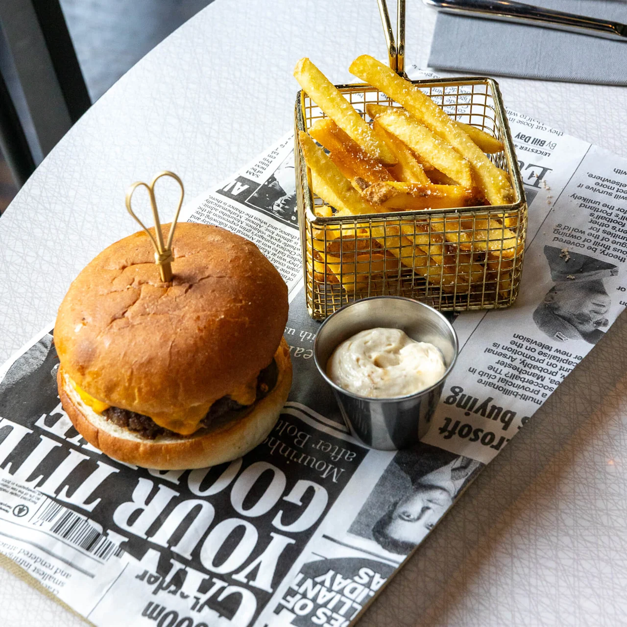 A burger with melted cheese sits on a newspaper-lined tray beside fries in a wire basket and a cup of sauce, on a white textured table. Text: "YOUR GOOD".
