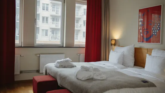 A neatly made bed with two robes lies in a modern hotel room. Three large windows reveal nearby apartment buildings. Red curtains and decor create a warm ambiance.