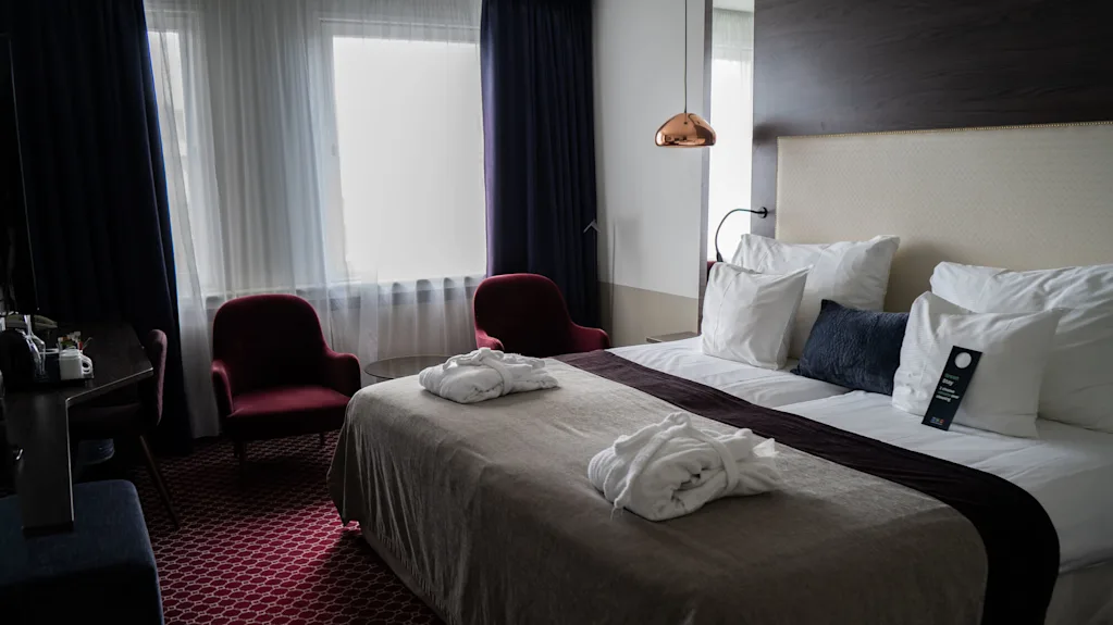 A neatly made hotel bed with robes and a sign on pillows, surrounded by two red chairs, a table, and large bright windows in a cozy room.
