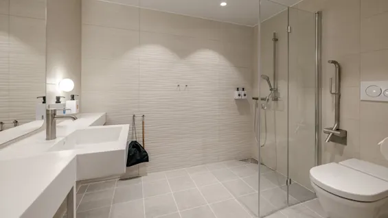 Et moderne bad har en veggmontert vask, en dusjkabin i glass og en toalett. Beige fliser og et stort speil skaper en romslig, elegant atmosfære.