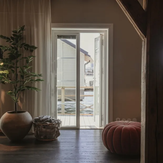 Et hyggeligt værelse med en stor potteplante og en rund puf, med udsigt gennem en åben dør til en vandudsigt med en bygning.