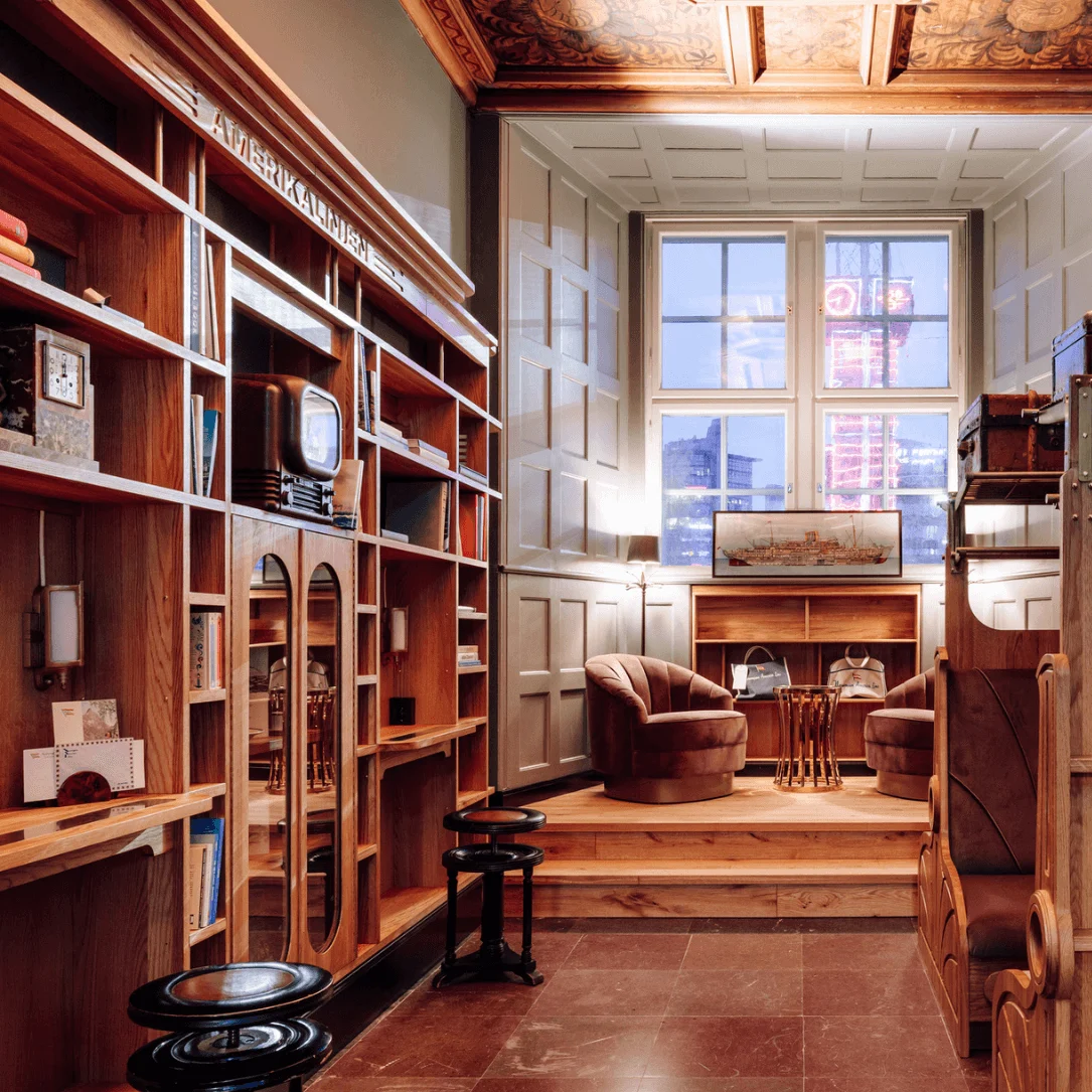 A cozy room features vintage suitcases on wooden shelves and a large window with city views. Two plush chairs sit around a small table. Text on shelves: "AMERIKALINIEN".