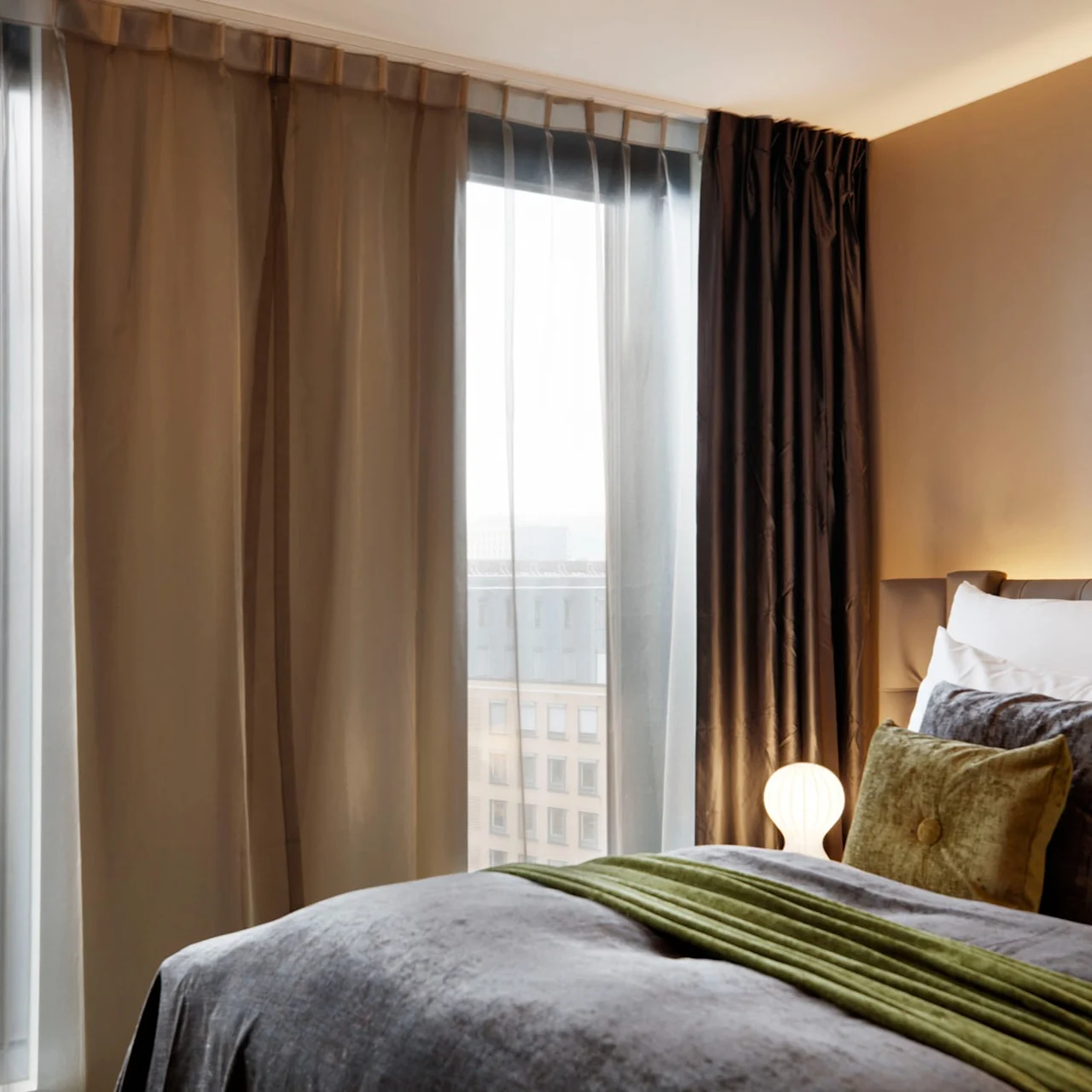 A bed with colorful pillows and a green throw lies by large windows under sheer curtains in a modern hotel room. Outside, tall buildings rise in the cityscape.