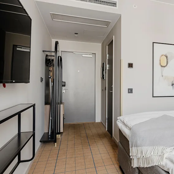 En prydligt bäddad hotellsäng med vita lakan och en grå filt står mot en brun vadderad sänggavel. Ett modernt, minimalistiskt rum med väggmonterad TV och abstrakt konst.