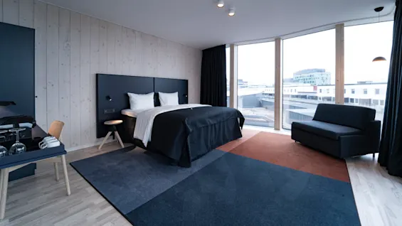 A modern hotel room features a neatly made double bed with black and white bedding, large windows overlooking city buildings, a sofa, and a small desk with a chair.