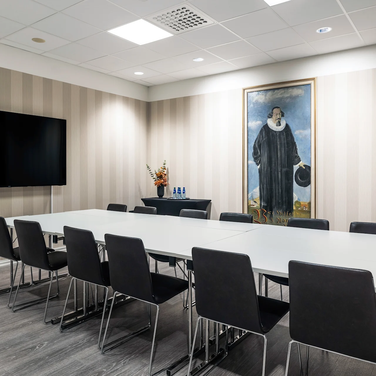Meeting room with a long white table surrounded by black chairs. A large framed painting hangs on striped wallpaper. A flat-screen TV is wall-mounted, and water bottles sit on a side table.
