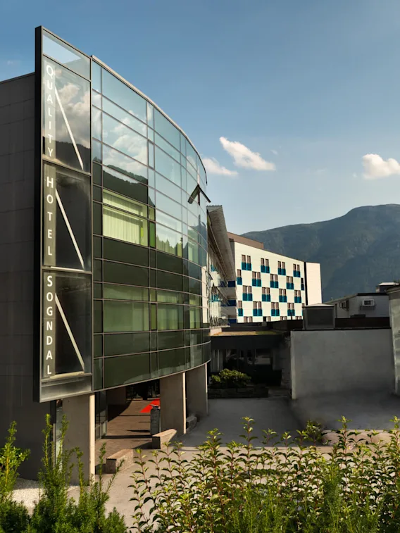 A modern glass building labeled "QUALITY HOTEL SOGNDAL" stands adjacent to traditional structures, set against a mountainous backdrop under a clear blue sky.