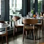 Et koselig restaurantinteriør med moderne brune stoler og runde bord dekket med glass, tallerkener, blomster og tente lys, ved store vinduer.