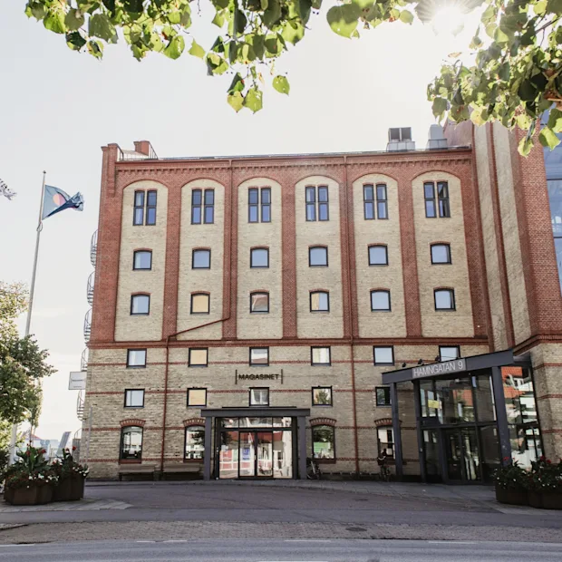 Suuri tiilirakennus, jossa modernit lasiosat ja sisäänkäynti. Liput ja puut kehystävät aurinkoista näkymää. MAGASINET, HAMNGATAN 9.