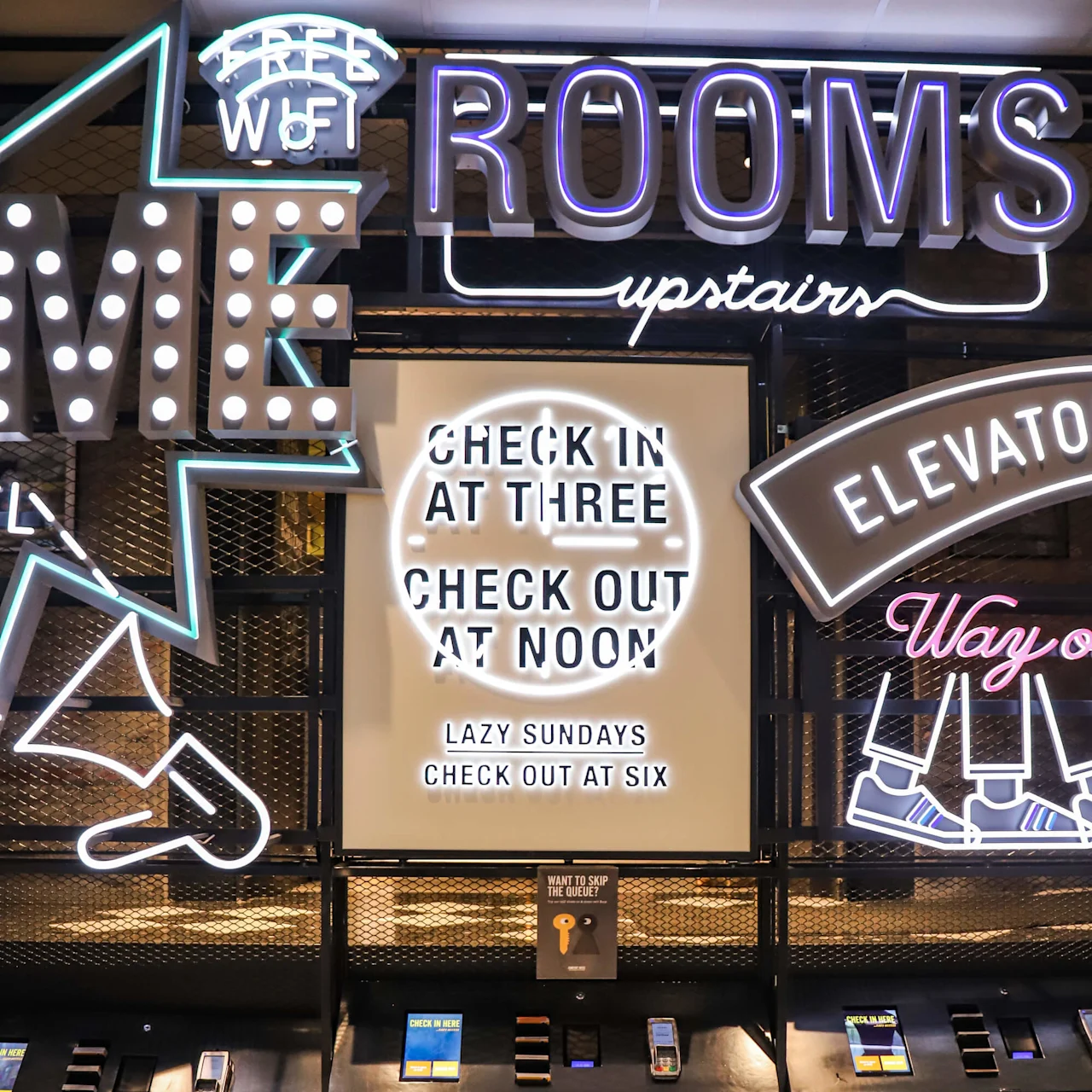 Neonskilt viser en hotellinnsjekking. Tekst: "CHECK IN AT THREE CHECK OUT AT NOON LAZY SUNDAYS CHECK OUT AT SIX." Omgivende ord inkluderer "ROOMS," "ELEVATOR," og "FREE WIFI."