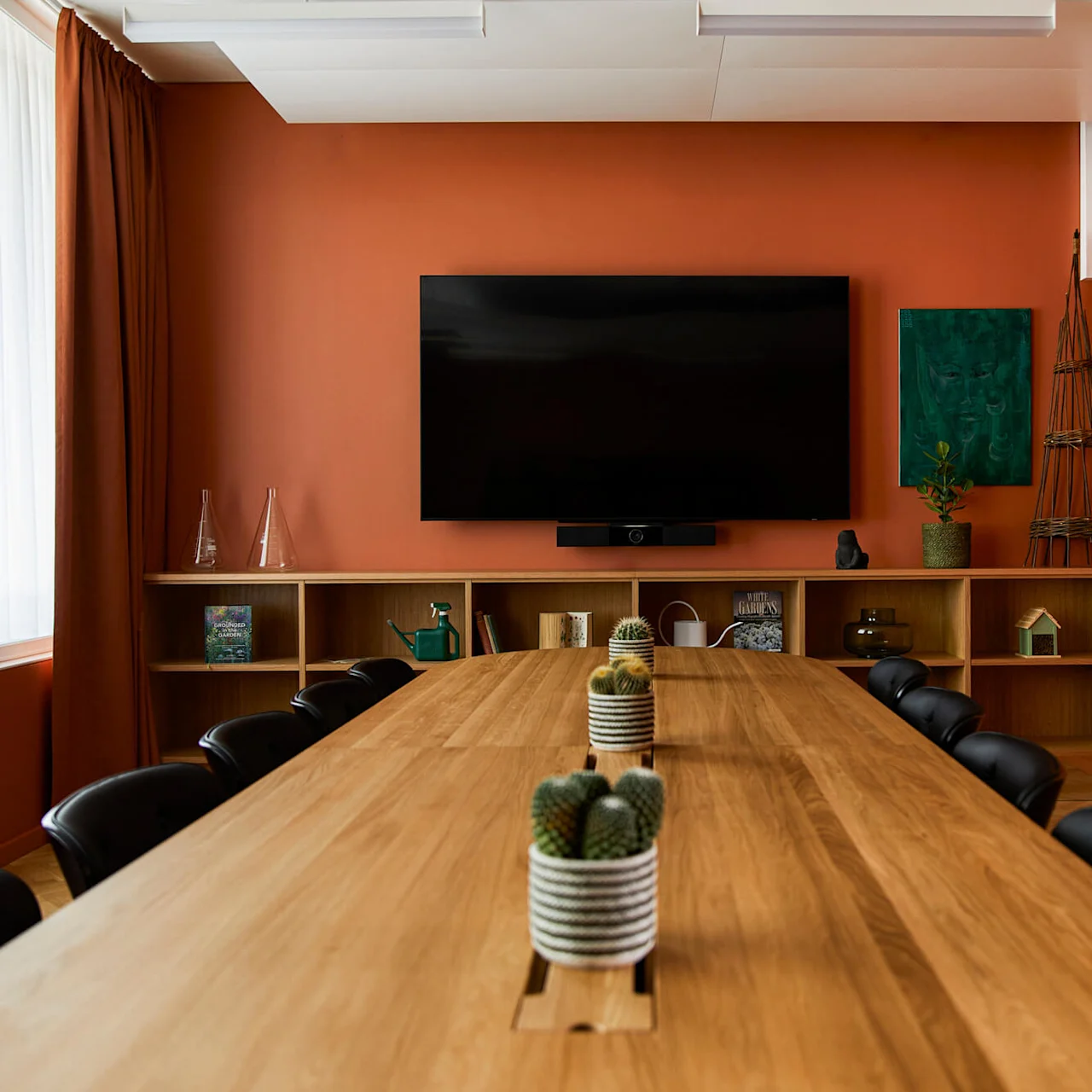 Et langt konferencebord i træ med potteplanter kaktusser i et rum med orange vægge, sorte stole, haveredskaber, hylder og et stort vægmonteret TV.