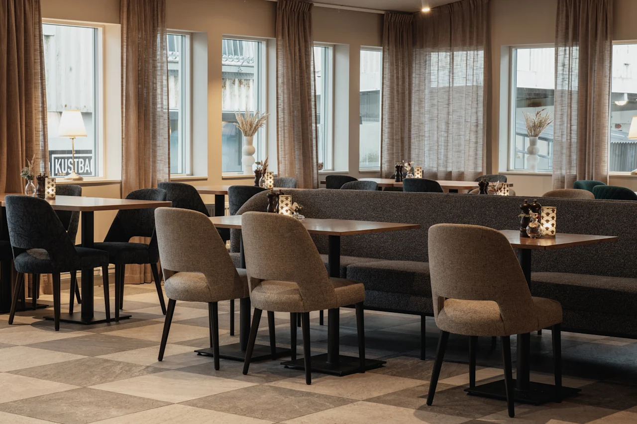 Et hyggeligt restaurantinteriør med borde, stole og bænkesæder, med store vinduer og et skilt, der læser KUSTBA.