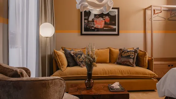 En senapsgul soffa med mönstrade kuddar står mot en beige vägg, under en abstrakt lampa, i ett mysigt rum. Ett träbord med en vas kompletterar närliggande stolar.