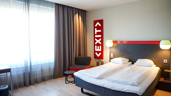 Sengen står pent i et moderne rom; stort vindu med gardiner slipper inn lys, rød "EXIT"-skilt henger på veggen ved siden av stol og lite bord.