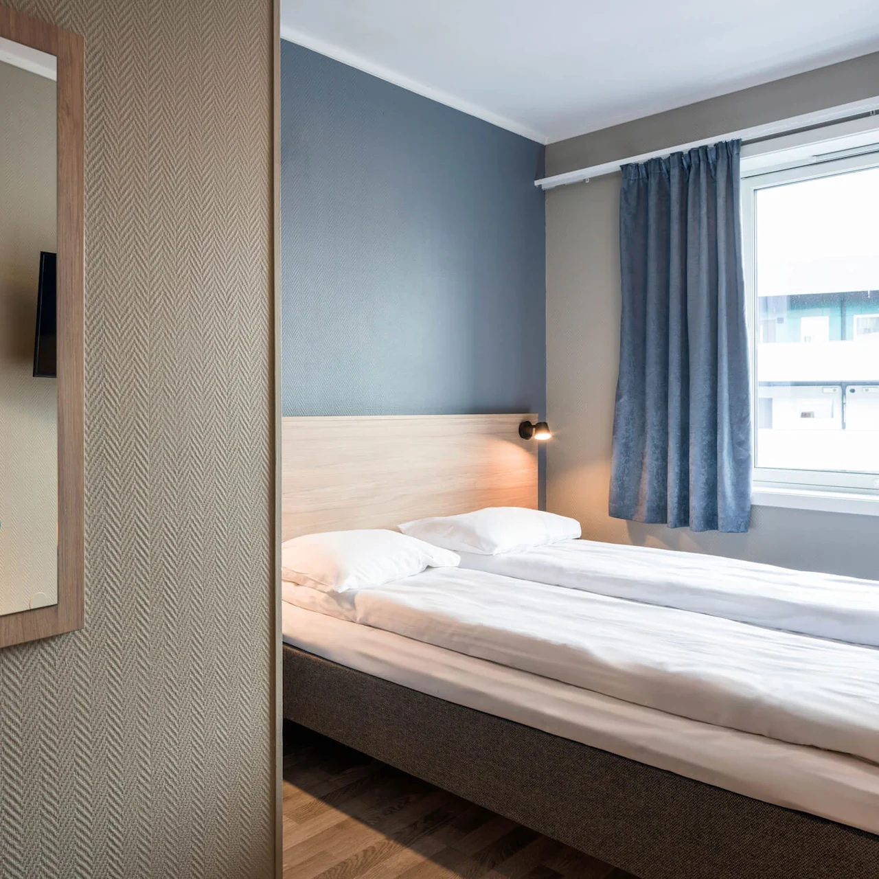 A bed with white sheets and pillows stands in a minimalistic room with wooden floors and a window with blue curtains; a mirror and lamp are visible on the left wall.