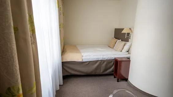 Et innbydende hotellrom med en komfortabel enkeltseng, et lite nattbord og dekorative gardiner.