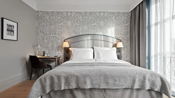 A cozy hotel room with a large bed, patterned wallpaper, and a desk. Sunlight streams through the window.