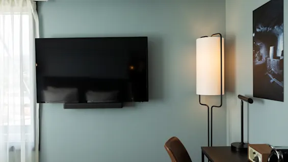 Et komfortabelt hotellrom med en veggmontert TV, en stilig gulvlampe og et skrivebord med en stol.