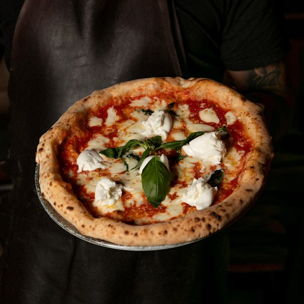 En person i forklæde holder en nybagt pizza toppet med tomat, mozzarella, burrata og basilikum. Teksten lyder CINO S Deli.