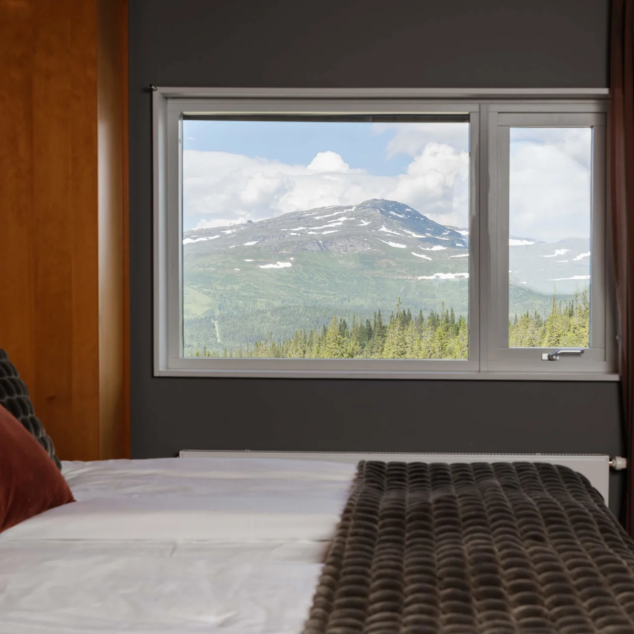 A bed with pillows faces a window showcasing a mountain view. The scene suggests a cozy interior with a nature-focused outlook.