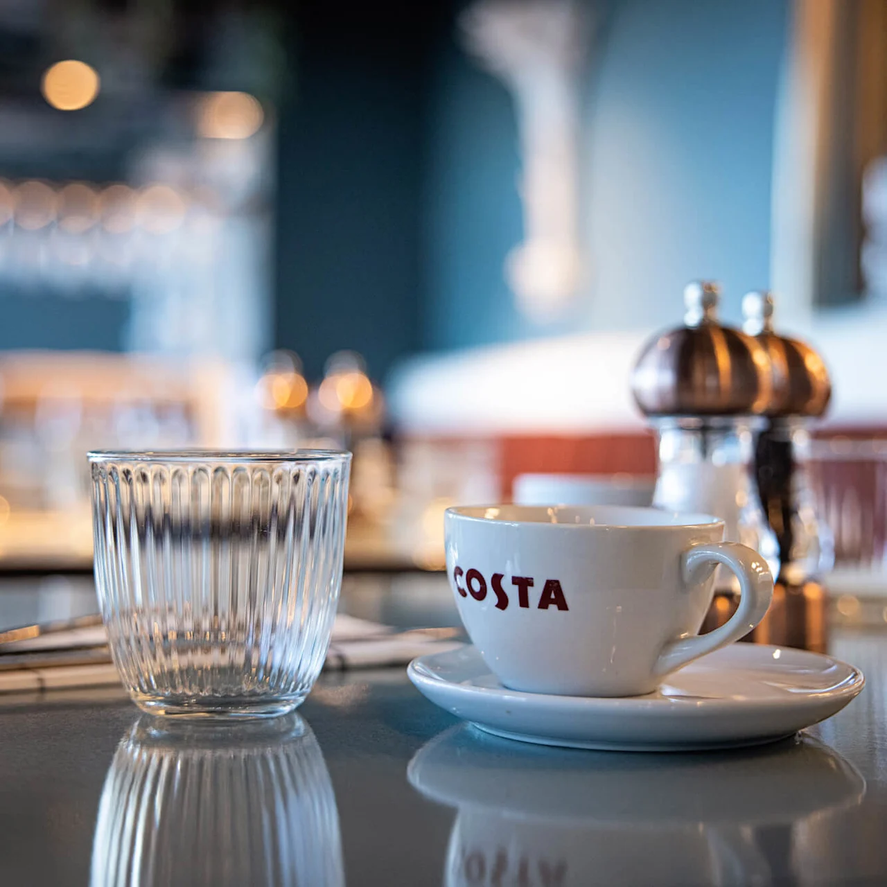 A white cup labeled "COSTA" sits on a saucer beside a clear glass and silverware in a softly lit cafe with blurred background details.