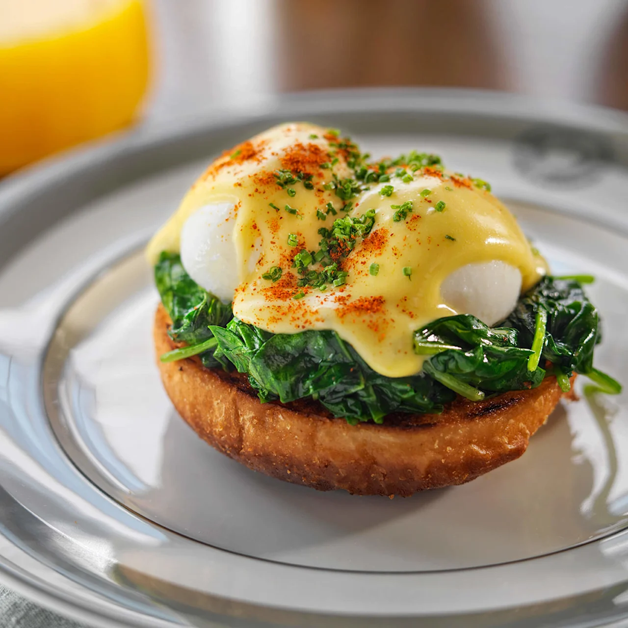 Eggs Benedict på ristet brød med spinat og hollandaise sauce, serveret på en tallerken med et glas appelsinjuice i baggrunden.
