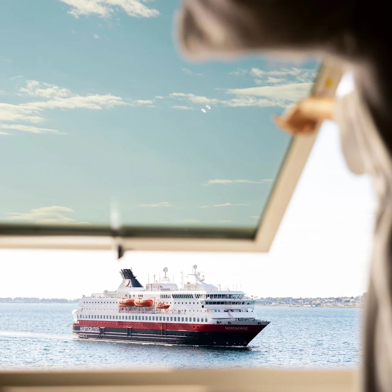 Et krydstogtskib mærket "NORDNORGE" sejler på roligt vand, set gennem et åbent vindue holdt af en person. Himlen er klar med spredte skyer.